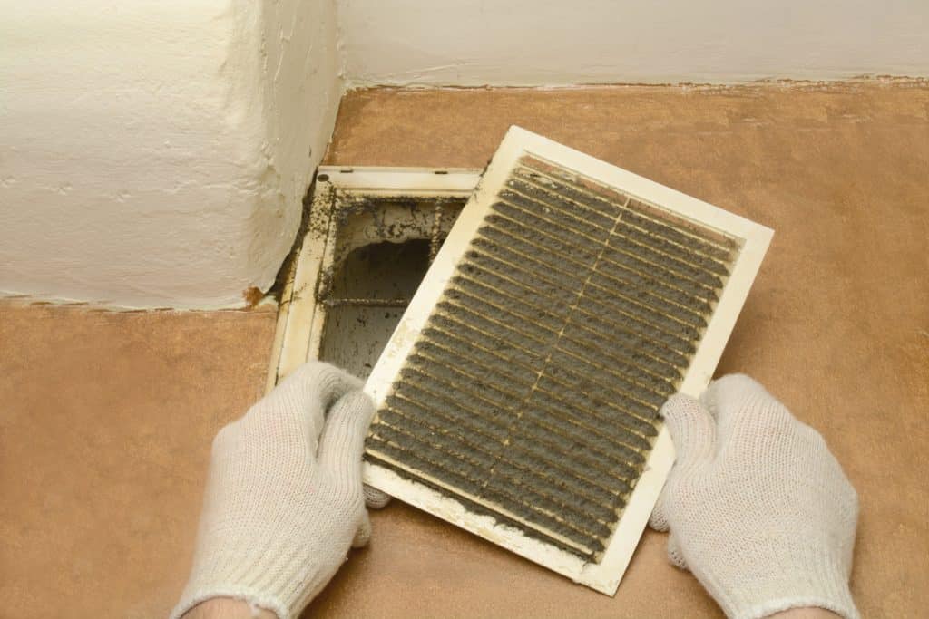 A person wearing gloves removes a heavily dust-covered ventilation grille from a wall, exposing a dirty air vent opening.